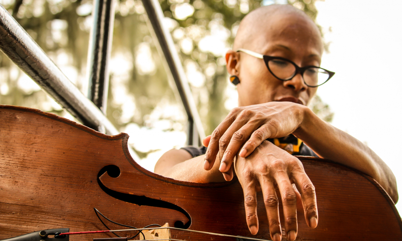 The musician Spirit McIntyre's portrait against a window. We see green in the background as the artist a brown skinned woman with black cats eye glasses looks down meditavely her hands draped across her instrument which is bass. 